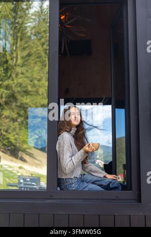 Junge Frau mit Kaffee am Fenster einer Waldhütte. Junge Frau hält eine Kaffeetasse, während sie an einem großen Hüttenfenster sitzt, mit Waldreflexionen. Stockfoto