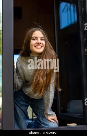 Lächelnde junge Frau am Kabinenfenster. Lächelnde junge Frau, die sich durch ein modernes Kabinenfenster in einer gemütlichen natürlichen Umgebung lehnt. Stockfoto