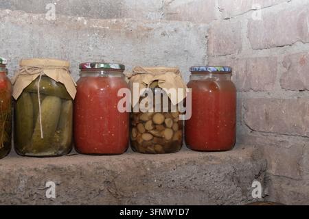 Verschiedene hausgemachte eingelegte Gemüsesorten, Gurken und Tomatensaft, die in Gläsern auf Regalen in einem rustikalen Keller gelagert werden. Stockfoto