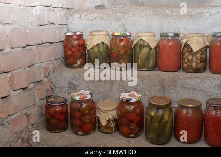Verschiedene hausgemachte eingelegte Gemüsesorten, Gurken und Tomatensaft, die in Gläsern auf Regalen in einem rustikalen Keller gelagert werden. Stockfoto