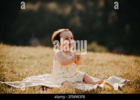Lachendes Baby-Mädchen sitzt auf einer Decke in einem sonnendurchfluteten Feld während der Goldenen Stunde Outdoor-Porträt Stockfoto