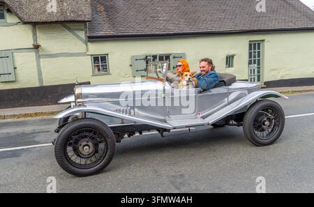 Ein Mann und eine Frau genießen eine malerische Fahrt in einem Alvis 12/50, während ihr Corgi mit ihnen vor einem traditionellen Strohdachhaus sitzt Stockfoto