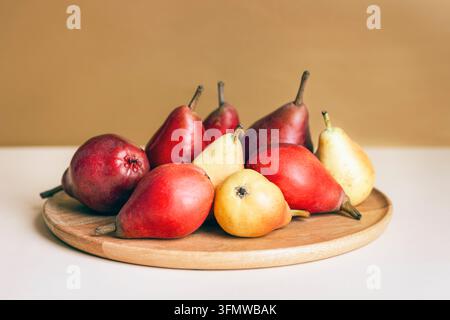 Rote und gelbe Birnen, die auf einem hölzernen Servierteller angeordnet sind, vor einem warmen beigen Hintergrund. Herbsterntekonzept, Nahaufnahme. Stockfoto