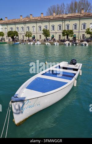 Blick auf ein weißes Boot namens Giulia, das auf den türkisfarbenen Gewässern des Gardasees vertäut ist, mit einem historischen Gebäude und kleinen Booten entlang des Ufers in Italien. Stockfoto