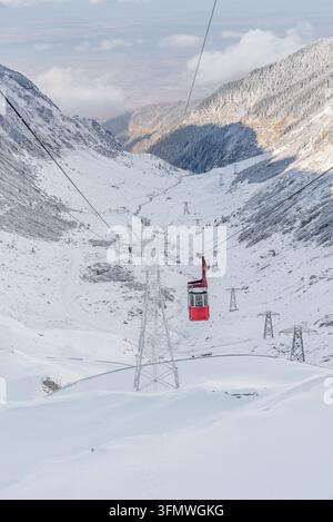 Blick auf eine rote Seilbahn, die über ein schneebedecktes Bergtal mit Stütztürmen entlang der Transfăgărășan in Curtea de Argeș, Județul Argeș, Stockfoto