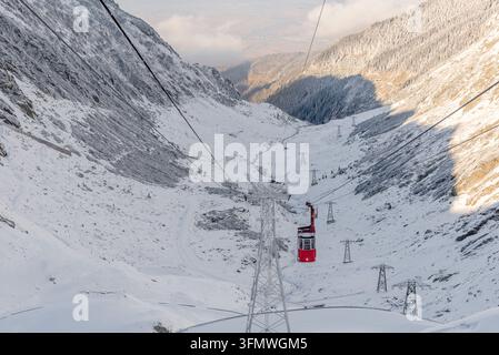 Blick auf eine rote Seilbahn, die über ein schneebedecktes Bergtal mit steilen Felshängen und Stützpfeilern entlang der Transfăgărăşana in Curtea de Stockfoto