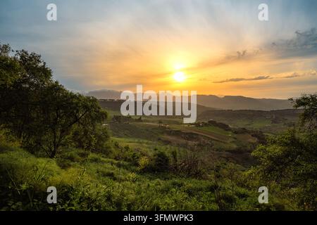 Die Sonne geht in einem glühend goldenen Himmel über geschichteten Bergkämmen unter und wirft warmes Licht auf grüne Felder, Olivenhaine und verstreute Bauernhäuser in der Stockfoto