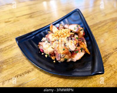 Thailändischer knuspriger Schweinebauch, gebraten mit Chilisalz, Knoblauch und Frühlingszwiebel, serviert auf einem schwarzen Teller als herzhafte, scharfe thailändische Restaurant-Speise. Stockfoto