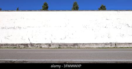 Verwitterter und geschälter weißer Putz an der umgebenden Wand am Straßenrand. Zweispurige Straße vorne, Himmel und Baumwipfel dahinter. Hintergrund für Kopierraum. Stockfoto