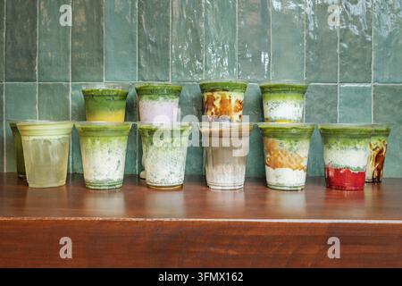 Verschiedene Matcha-Getränke in zwei Reihen von Tassen, die auf einer hölzernen Theke mit gefliester Wand präsentiert werden Stockfoto