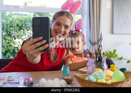 Mutter und Kind in Hasenohren, die mit Telefon und Pinsel Ostereier mit Farben bemalen zu Hause Stockfoto