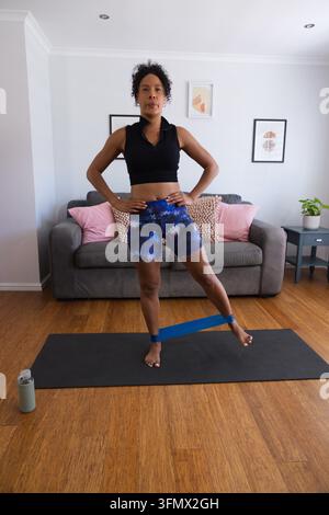 Afroamerikanische Frau, die mit einem blauen Loop-Band auf einer Matte zu Hause im Sportoutfit einbeinigen Gleichgewichtstest durchführt Stockfoto