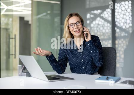 Professionelle Geschäftsfrau sitzt am Bürotisch, führt ein Telefongespräch, lächelt und gestikuliert, kommuniziert mit Kunden oder Kollegen während des Arbeitstages, zeigt einen glücklichen und selbstbewussten Ausdruck Stockfoto
