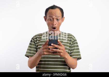 Überraschter asiatischer Mann in grünem Hemd, der sein Smartphone benutzt, isoliert vor weißem Hintergrund. das Konzept, von Informationen überrascht zu sein, die man erfährt thro Stockfoto