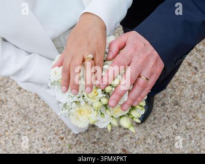Nahaufnahme der Hände einer Braut und eines Bräutigams mit Eheringen auf einem weißen Blumenstrauß. Stockfoto
