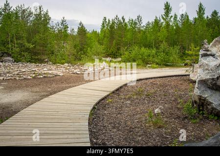 Gekrümmter Holzsteg windet sich durch Kies und grünen Wald. Der Weg lädt zur ruhigen Erkundung der natürlichen Landschaft ein. Ideal für Natur, Wandern oder Wohlbefinden Stockfoto