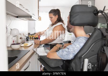 Eine Frau bereitet in einer Küche Essen und Getränke zu, während sie einem Mann im Rollstuhl hilft. Sie teilen den Raum in einem barrierefreien Zuhause und arbeiten zusammen. Stockfoto