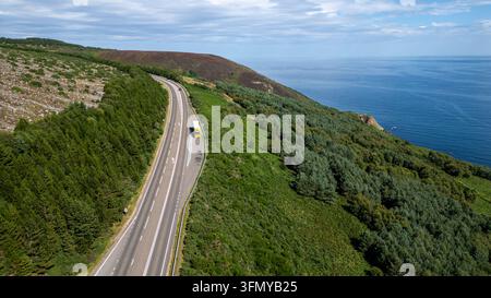 Eine Luftaufnahme zeigt eine Küstenstraße, die sich durch eine üppige grüne Landschaft windet, mit dem tiefblauen Ozean im Hintergrund unter einem bewölkten Himmel in Schottland Stockfoto