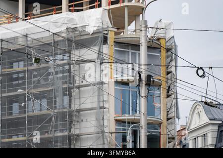 Überhead-Verkehrsleitungen kreuzen einen Strommast neben einem gerüsteten Gebäude mit freiliegender Fassadenisolierung, was städtische Bauschnittsinterfaces veranschaulicht Stockfoto