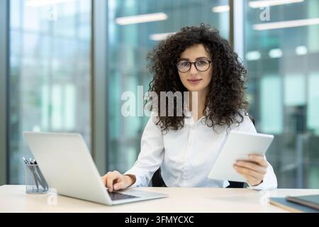 Geschäftsfrau mit lockigem Haar, die eine Brille und ein weißes Hemd trägt, einen Laptop benutzt und ein digitales Tablet hält, und in einer modernen Firmenumgebung in die Kamera schaut Stockfoto