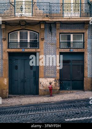 Historische Wohnhausfassade in Lisboa mit kunstvollen schmiedeeisernen Balkonen, gemusterten Azulejo-Fliesen, dunklen Türen, Kopfsteinpflaster und einem roten Fi Stockfoto