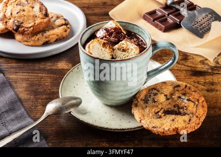 Gemütliche Tasse heiße Schokolade mit gerösteten Marshmallows und Kakaopulver, serviert mit Schokoladenkeksen und dunkler Schokolade auf rustikalem Holz Stockfoto