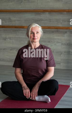 Reife Frau sitzt im Schneidersitz auf einer roten Yogamatte in einem ruhigen Indoor-Studio und schaut in die Kamera. Porträt mit Kopierraum betont Wellness, Flexibi Stockfoto