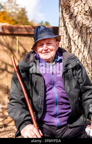 Älterer Bauer mit dunklem Hut und Jacke ruht neben einem großen Baum, hält einen Holzstock in einem ländlichen Hof an einem sonnigen Herbsttag. Stockfoto