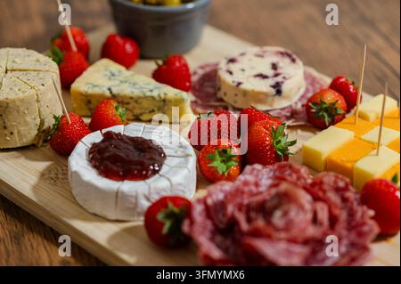 Holzservierbrett mit sortiertem Käse, luftgetrocknetem Fleisch, frischen Erdbeeren, Marmelade und Zahnstochern für einen appetitlichen Party-Snack, eine Vorspeise oder ein Gougerie Stockfoto
