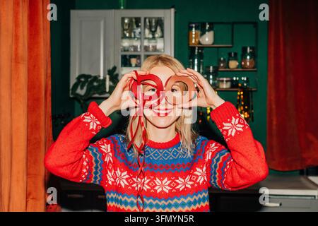 Lächelnde Frau in einem farbenfrohen festlichen Pullover hält ein rotes Geschenkband über ihren Augen wie eine Brille, während sie in einer gemütlichen, weihnachtlich geschmückten Küche posiert. Stockfoto