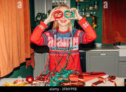 Lächelnde Frau in einem festlichen Pullover hält rote und grüne Bänder wie eine Brille, während sie auf einem Küchentisch verpackte Weihnachtsgeschenke und Dekorationen vorbereitet Stockfoto