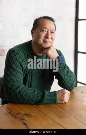 Reifer asiatischer Mann sitzt an einem Holztisch nahe dem Fenster, trägt einen grünen Kabelpullover und eine Uhr, das Kinn ruht auf der Tischfläche. Porträt, Lifestyle, Strickware, Smaragd, Holzmaserung Stockfoto