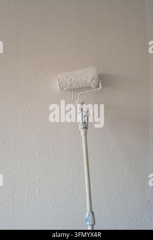 Nahaufnahme eines Farbrollers, der mit weißer Farbe bedeckt ist und vor einer texturierten Innenwand steht, zur Veranschaulichung von Hausrenovierung, Hausumbau und professioneller P Stockfoto