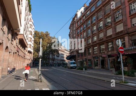 STRASBOURG, FRANKREICH - 21. SEPTEMBER 2024: Rückansicht einer Frau in rotem T-Shirt, die entlang einer Stadtstraße mit historischen Gebäuden und Straßenbahnschienen läuft Stockfoto