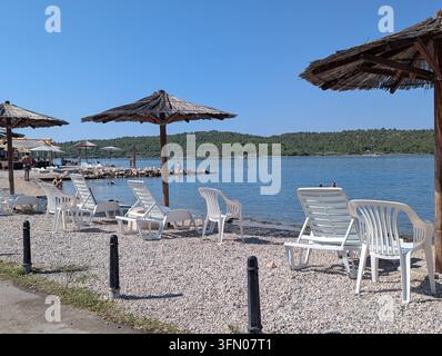Pirovac, Kroatien, 16. August 2026 Leere weiße Loungestühle und Strohsonnenbeschattungen säumen einen mit Kies bedeckten Uferstreifen neben ruhigem blauem Wasser, mit Strand, Meeresufer, Ufer Stockfoto