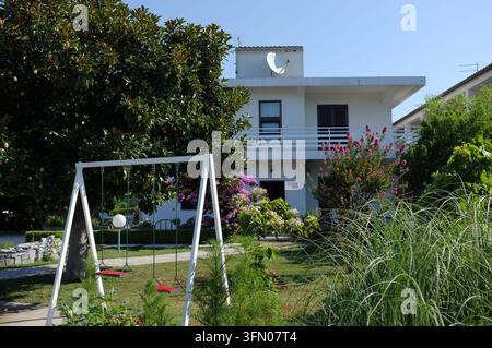 Pirovac, Kroatien, 16. August 2026 Helles Wohngebäude mit Balkon, Satellitenschüssel, Gartenschaukel und üppiger Bepflanzung unter klarem blauem Himmel, mit h Stockfoto