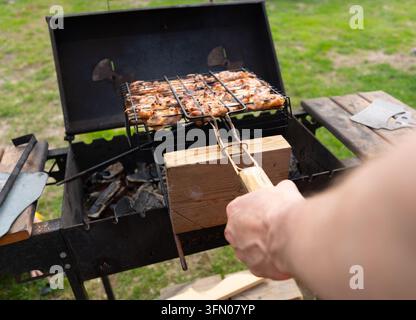 Ein Mann hält einen Fleischspieß über einem Grill und präsentiert handwerkliche Grillkunst in einer Nahaufnahme. Der konzentrierte Koch grillt gekonnt Kebab in einem sonnigen Hinterhof mit sicherer Präzision. Stockfoto