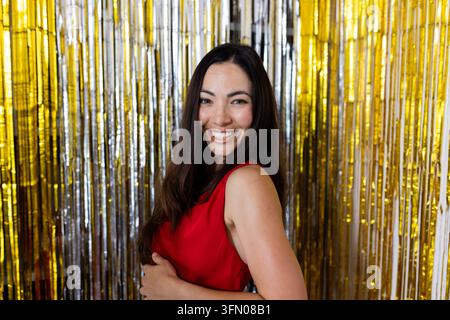 Asiatische Frau lächelt und posiert in rotem ärmellosem Kleid an einem Eventstand mit Lametta-Vorhang Stockfoto