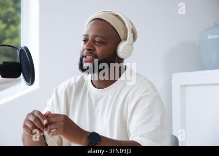 Afroamerikanischer Mann spricht am Tisch im Studio mit Mikrofon, trägt Kopfhörer und Beanie Stockfoto