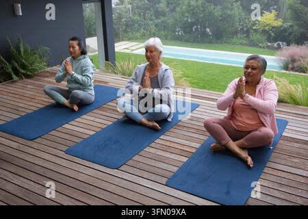 Verschiedene Freundinnen sitzen und drücken die Handflächen auf drei blauen Yogamatten auf einem Holzdeck in der Nähe des Pools Stockfoto