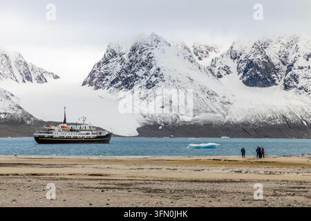Expeditionsschiff und Touristen an einem abgelegenen arktischen Strand mit treibendem Eisberg und dramatischen schneebedeckten Bergen im Sommer, Spitzbergen, Svalbard Stockfoto