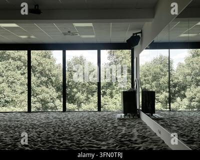 Frankfurt am Main, Deutschland - 30. Juni 2026: Sandbedeckter Fitnessbereich im Innenraum mit großen Fenstern, die auf Bäume hinausgehen, und einer verspiegelten Wand. Stockfoto
