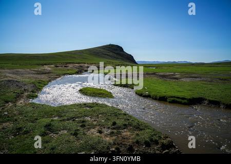 Ein schmaler, gewundener Fluss mit glitzerndem Wasser fließt durch eine lebendige grüne Steppenlandschaft, umgeben von sanften Hügeln unter einem klaren blauen Himmel. Ein klei Stockfoto