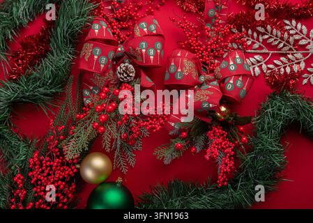 Überkopf-Studioaufnahme zeigt Weihnachtsgirlande, rote Schleifen mit Schriftzug, Tannenzapfen, rote Beeren, Christbaumkugeln und Lametta vor lebhaftem rotem Hintergrund Stockfoto