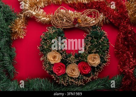 Hell beleuchtete Studioaufnahme, die einen festlichen Weihnachtskranz zeigt, der mit Kunstblumen dekoriert und mit einer Juteschleife gebunden ist, umrahmt von funkelndem Gold, Rot und Grün Stockfoto