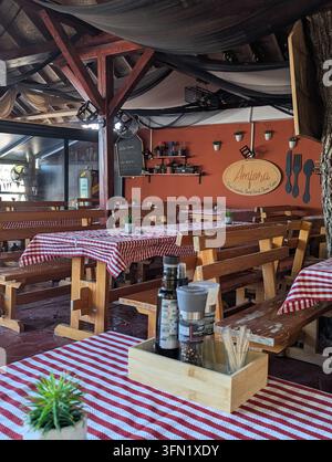 Pirovac, Kroatien, 16. August 2026 Rustikales Open-Air-Restaurant-Interieur mit Holzbänken, karierten Tischdecken, hängender Dekoration, Terracotta-Wand Stockfoto