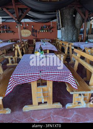 Pirovac, Kroatien, 16. August 2026 Leerer rustikaler Essbereich mit Holzbanken, karierten Tischdecken, Topfdekoration, Terracotta-Wänden, hängendem Stoff Stockfoto