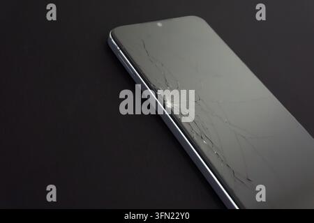 Rissiger Smartphone-Bildschirm mit radial zerbrochenem Glas vom Aufprallpunkt, auf dunkler Oberfläche mit Kopierraum Stockfoto