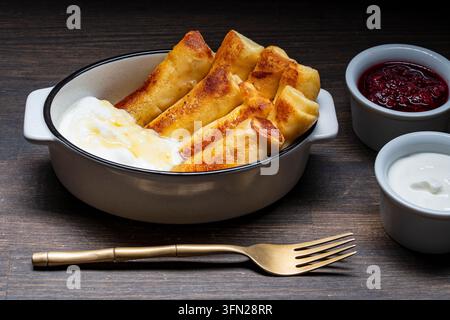 Leckere gebratene Crêpes gefüllt mit Hüttenkäse, serviert mit süßem Honig, saurer Sahne und roter Beerenmarmelade in einer rustikalen Schüssel auf dunklem Holzgrund. Per Stockfoto