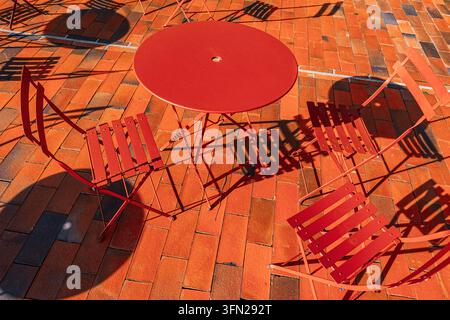 Rote Metall-Café-Stühle und -Tische werfen kühne geometrische Schatten über orangefarbene Ziegelpflaster im hellen Sonnenlicht, ohne dass Menschen anwesend sind Stockfoto